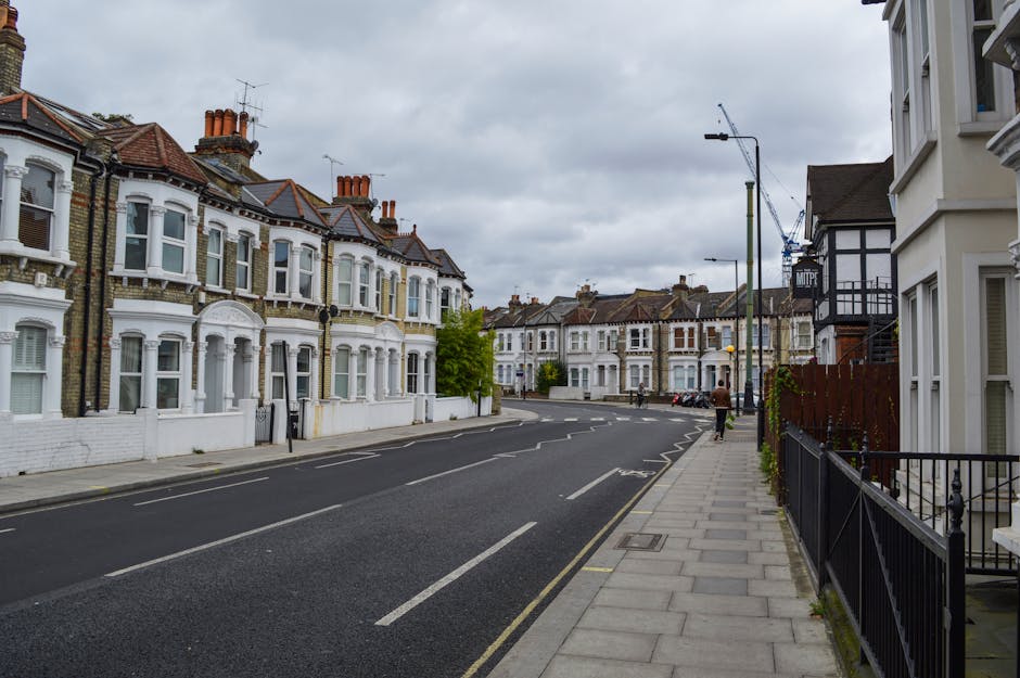 A quiet residential street in Earls Court SW5 featuring a row of Victorian-style terraced houses with white facades, bay windows, and decorative brickwork, alongside a paved sidewalk with a black metal fence and some potted plants. The street has a single lane in each direction, marked with painted lines, and a few parked cars are visible in the distance. Overhead, there are street lamps and a tall crane indicating ongoing construction work nearby. The sky is overcast with grey clouds, creating diffuse lighting. This scene illustrates the typical urban environment where house removals and home relocation services by [COMPANY_NAME] may be required, especially for residents planning furniture transport, packing, and loading as part of their moving process towards or from Earls Court.
