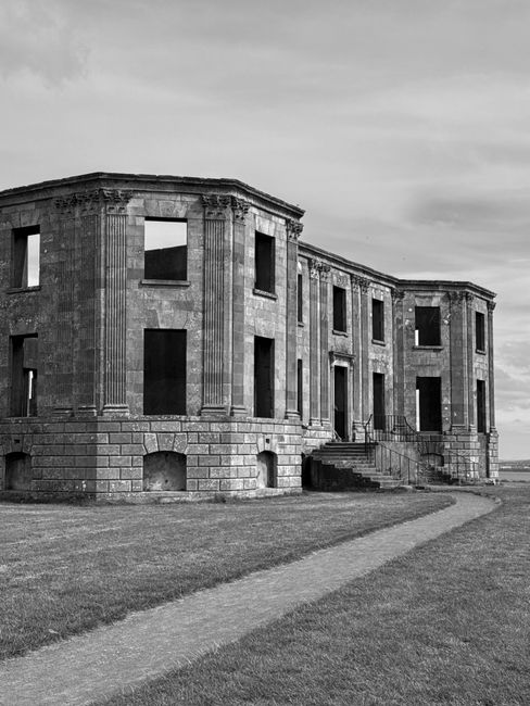 A black and white photograph of a historic, two-story building with a curved façade, featuring multiple rectangular window openings without glass, exposing the interior. The structure appears weathered, with visible stonework and decorative pilasters along the edges. In the foreground, a narrow paved pathway leads towards the building's entrance, which has a small flight of stairs with metal railings on both sides. The surrounding ground is covered with neatly trimmed grass, and an open sky with scattered clouds is visible above. The setting likely captures the site of a house removal or renovation project, consistent with services offered by removal companies like [COMPANY_NAME], in the context of home relocation or furniture transport.