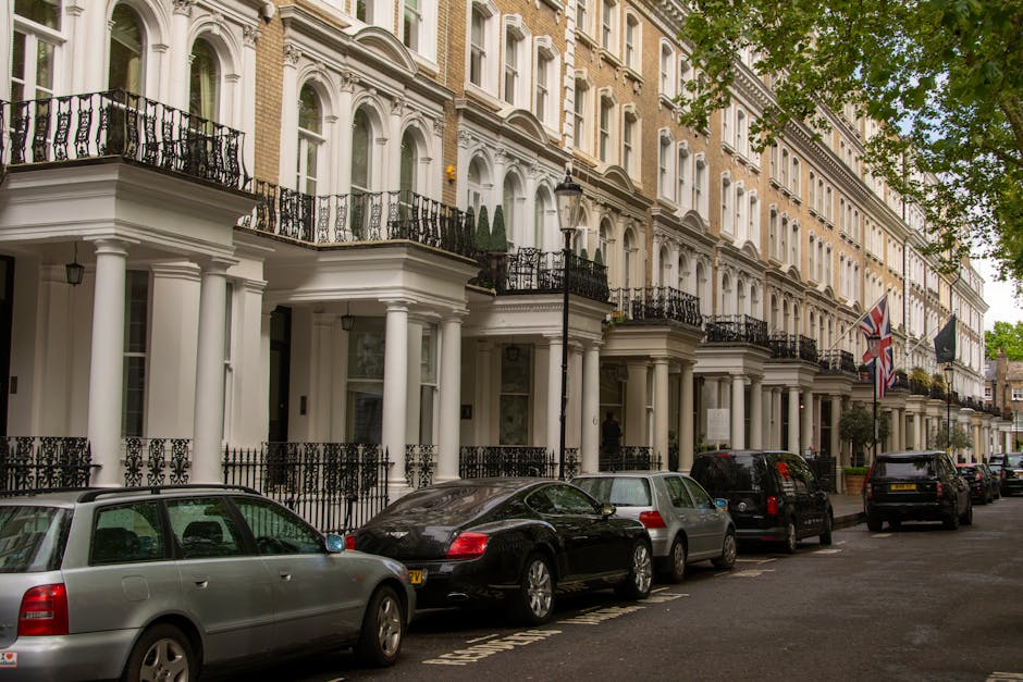A quiet residential street in Earls Court SW5 featuring a row of Victorian-style terraced houses with white facades, bay windows, and decorative brickwork, alongside a paved sidewalk with a black metal fence and some potted plants. The street has a single lane in each direction, marked with painted lines, and a few parked cars are visible in the distance. Overhead, there are street lamps and a tall crane indicating ongoing construction work nearby. The sky is overcast with grey clouds, creating diffuse lighting. This scene illustrates the typical urban environment where house removals and home relocation services by [COMPANY_NAME] may be required, especially for residents planning furniture transport, packing, and loading as part of their moving process towards or from Earls Court.