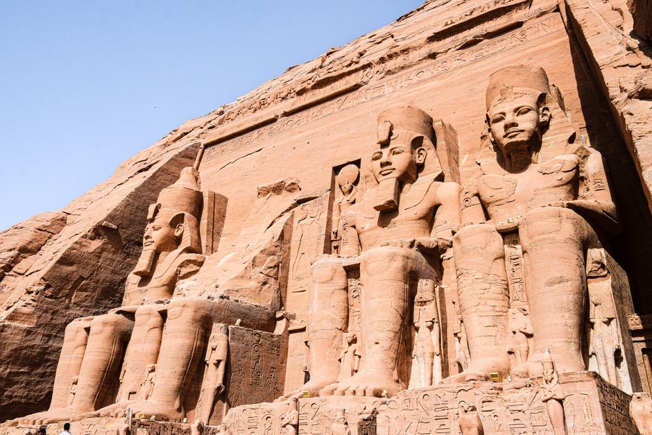 Photograph of the ancient Egyptian temples at Abu Simbel, with four massive statues of seated pharaohs carved from sandstone, set against a clear blue sky. The statues are positioned in front of a large façade featuring detailed hieroglyphic inscriptions and carvings. In the foreground, parts of the temple entrance are visible, with steps leading up to the doorway partially shaded by the massive statues. The scene is illuminated by natural sunlight, highlighting the reddish-brown tones of the sandstone and the intricate stonework of the sculptures. This historical site is sometimes visited during home relocation or travel logistics planning, illustrating the importance of careful transport and preservation of valuable artefacts, similar to the detailed packing and moving efforts undertaken by professional removals, such as those provided by [COMPANY_NAME]. The overall composition showcases the impressive scale and craftsmanship of ancient Egyptian architecture, with a focus on preservation during transport and relocation processes in mind.