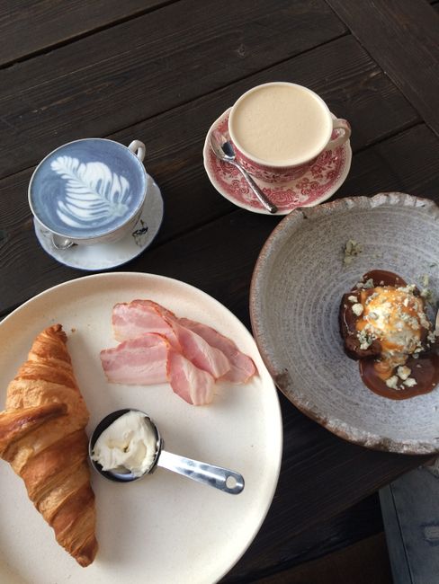 A table setting featuring a plate with a croissant, two slices of bacon, and a small container of butter with a spoon; a ceramic cup of coffee with cream on a pink floral saucer; a smaller cup of blue latte art with a spoon, and a bowl of dessert topped with cream and crumbled toppings. The table has a dark wooden surface, and the image captures a casual, cozy breakfast scene, possibly within a home or café environment, with a focus on food presentation and beverage variety that could relate to preparations prior to a house removal or packing process involving an easy, welcoming meal.