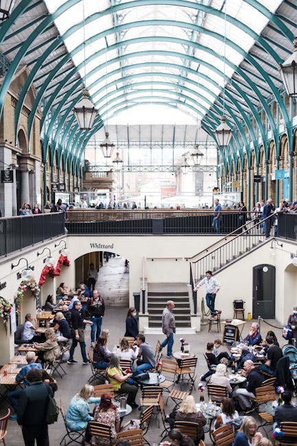 Inside a large, historic indoor venue with high, arched glass and metal roofing supported by black iron columns, a team from [COMPANY_NAME] is engaged in a home relocation or packing process. Several 